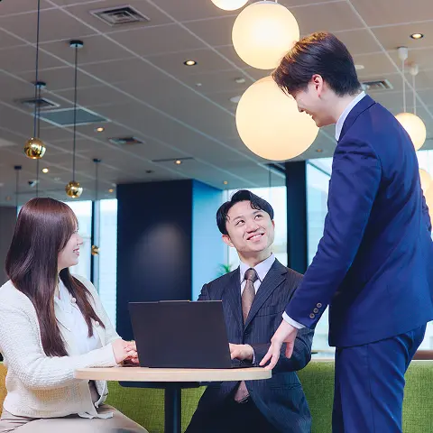 オフィスのラウンドテーブルを囲み、ノートパソコンを前に談笑しながら打ち合わせするビジネスパーソン3名。緑のソファと球形の照明がある明るい執務空間