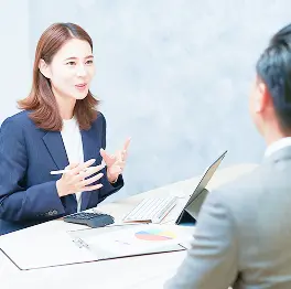 俯瞰で撮影したデスク越しの打ち合わせ。紺のスーツの女性が手を動かしながら微笑み、向かいのグレーのスーツの男性と話している。机にはタブレット、電卓、円グラフの資料