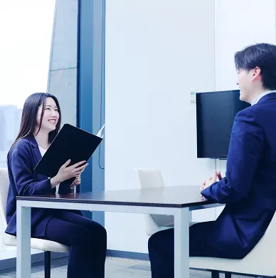 明るい会議室で対面に座る男女。女性は黒い書類フォルダを手に男性に向かって微笑み、男性は後ろ姿寄りで女性を見ている。窓の外にビル街、白い壁にはモニターが掛かっている
