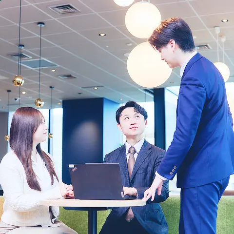 オフィスでスーツを着た男性2名と女性1名がPCを見ながら談笑している