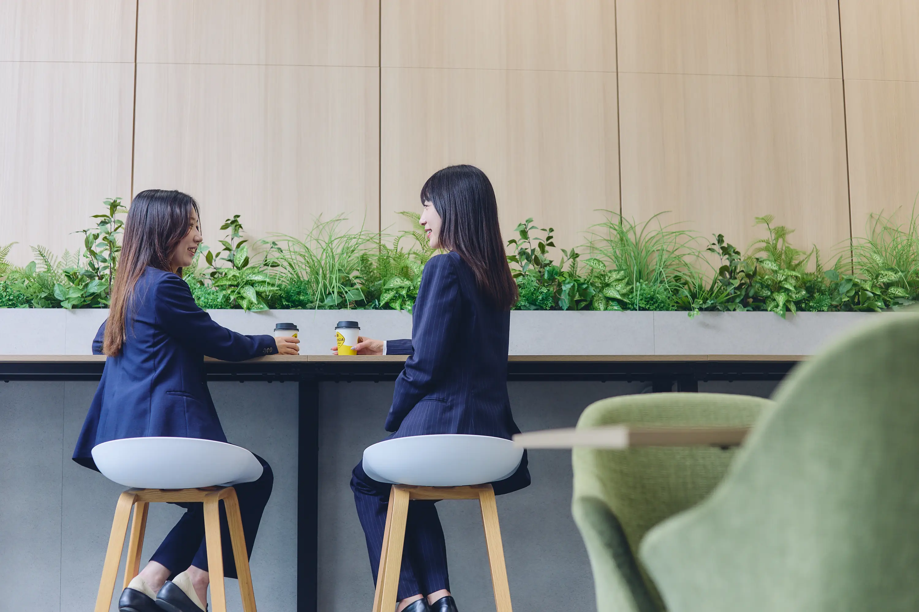 若い女性2名がコーヒーを手にオフィスで談笑している様子。