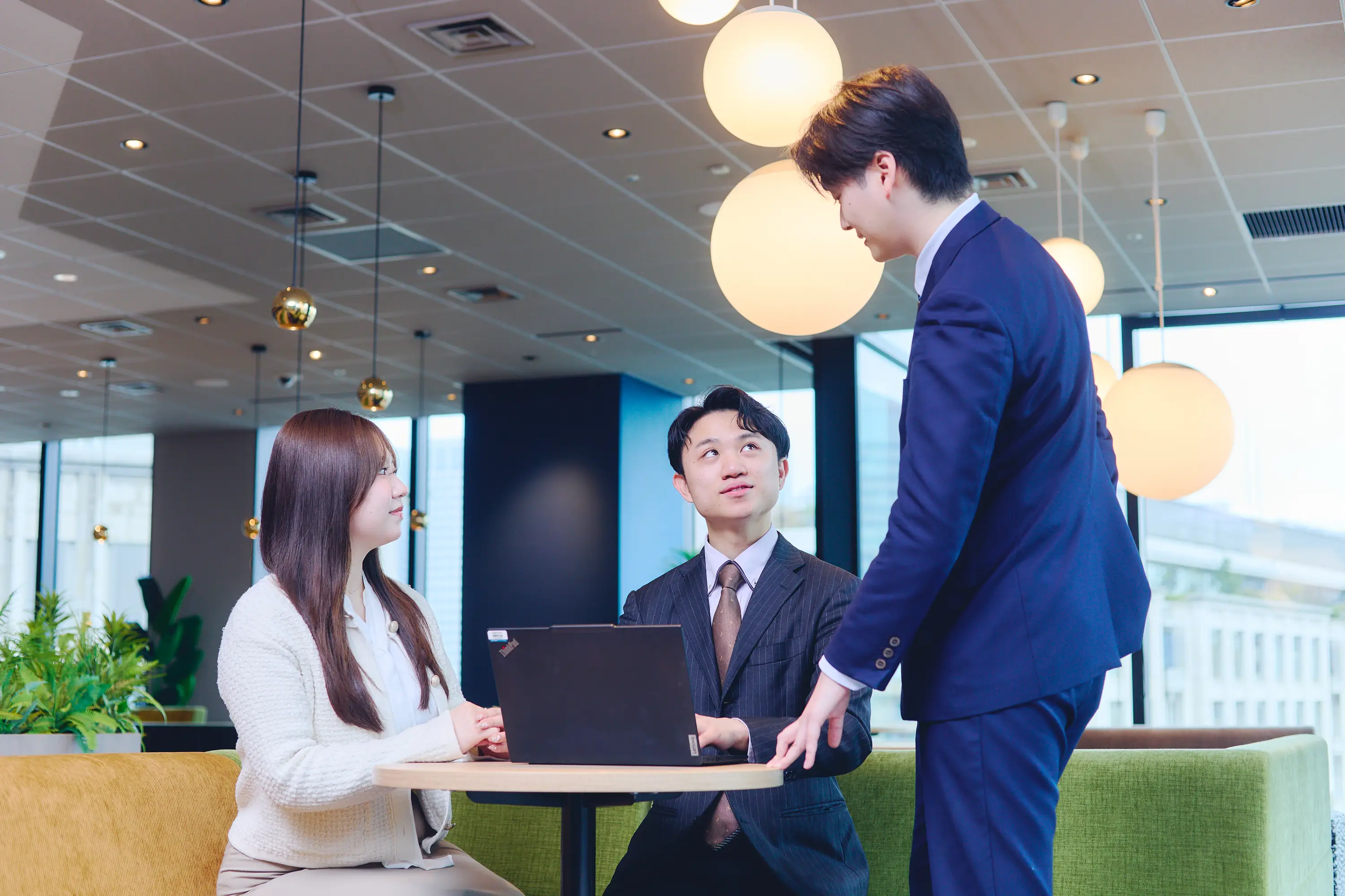 若い女性1名と男性2名がオフィスの歓談スペースでPCを囲みながら談笑している様子